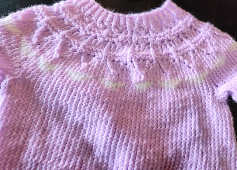 A short-sleeved, light purple knitted sweater featuring a textured yoke with a decorative pattern around the neckline. The garment is constructed with a ribbed collar, ribbed sleeve cuffs, and a ribbed hem, showcasing a consistent knit stitch throughout the body.