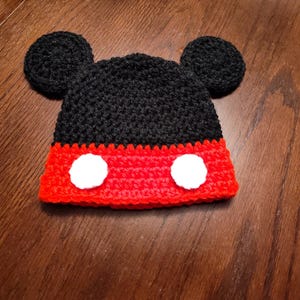 A black and red crocheted hat featuring two round black ears on top and two circular white patches on the front red section. The hat is constructed with a textured yarn stitch pattern, creating a distinct character-inspired design.