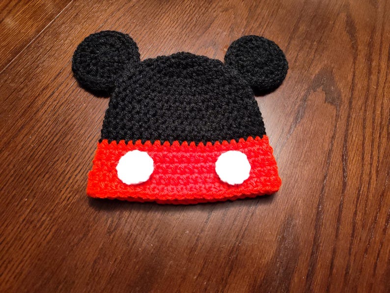 A black and red crocheted hat featuring two round black ears on top and two circular white patches on the front red section. The hat is constructed with a textured yarn stitch pattern, creating a distinct character-inspired design.