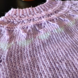 A light purple knitted sweater featuring a ribbed crew neckline and a textured yoke pattern. The yoke includes a horizontal band of white yarn integrated into the knit, creating a subtle contrast against the solid lavender color.
