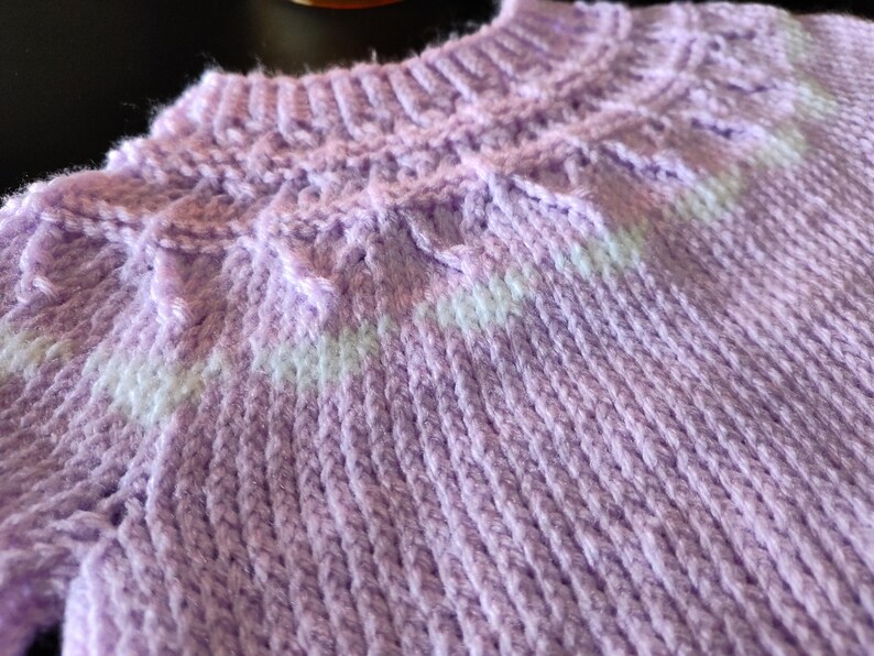 A light purple knitted sweater featuring a ribbed crew neckline and a textured yoke pattern. The yoke includes a horizontal band of white yarn integrated into the knit, creating a subtle contrast against the solid lavender color.