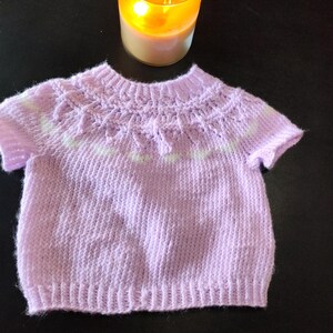 A short-sleeved, light purple knitted sweater featuring a ribbed crew neckline, a textured yoke with a subtle white horizontal stripe pattern, and a ribbed hem. The garment is constructed with a soft yarn texture and short sleeves.