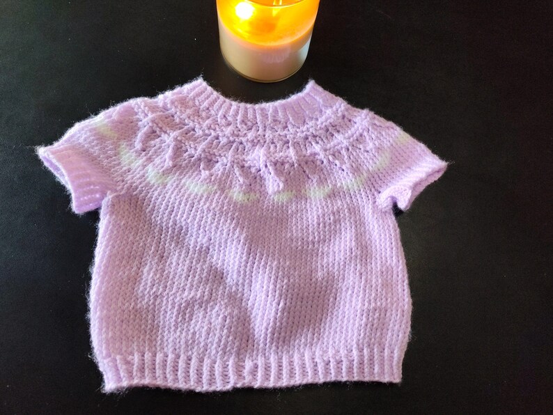 A short-sleeved, light purple knitted sweater featuring a ribbed crew neckline, a textured yoke with a subtle white horizontal stripe pattern, and a ribbed hem. The garment is constructed with a soft yarn texture and short sleeves.