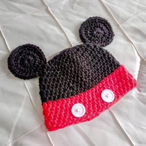 A crocheted beanie featuring a black upper section with two large circular ears attached to the sides. The lower portion is bright red and includes two white circular buttons stitched horizontally across the front of the hat.