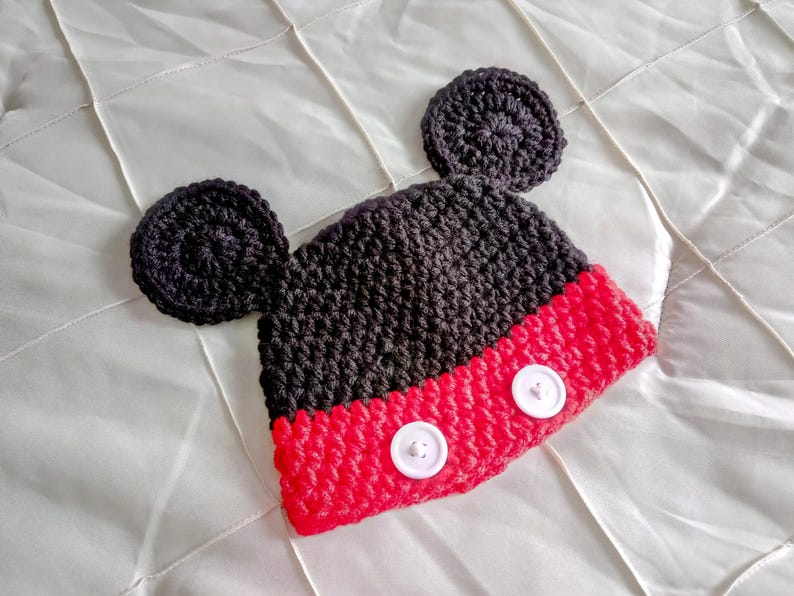A crocheted beanie featuring a black upper section with two large circular ears attached to the sides. The lower portion is bright red and includes two white circular buttons stitched horizontally across the front of the hat.