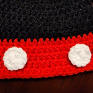 A handmade crochet hat featuring a black upper section and a vibrant red brim. Two circular white crochet appliques are attached to the front of the red brim, creating a distinct contrast against the bright red yarn texture.