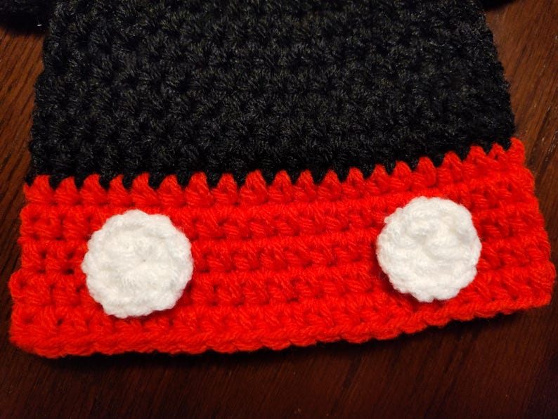 A handmade crochet hat featuring a black upper section and a vibrant red brim. Two circular white crochet appliques are attached to the front of the red brim, creating a distinct contrast against the bright red yarn texture.
