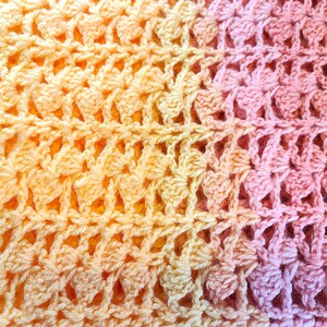 A handmade crochet blanket in shades of yellows, pinks, and purples. The blanket features a textured pattern and a scalloped edge.