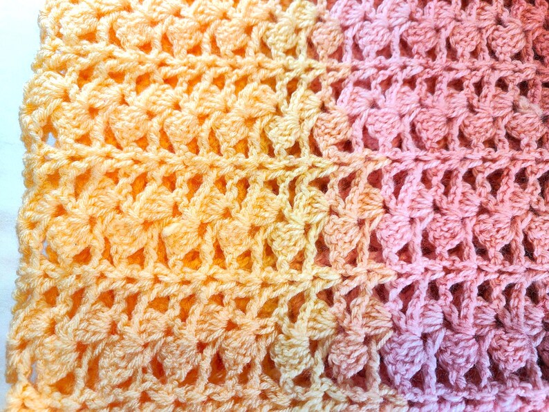 A handmade crochet blanket in shades of yellows, pinks, and purples. The blanket features a textured pattern and a scalloped edge.