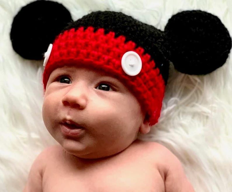 A red and black crocheted hat featuring two large round black ears on the sides and two white circular buttons attached to the front red band. The hat is constructed with a textured yarn stitch pattern and has a rounded crown shape.