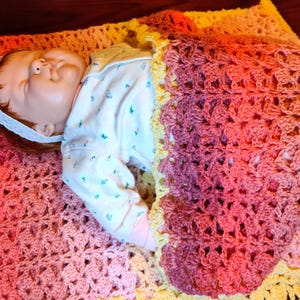 A doll is laying on it's back on a multi-colored striped blanket and is covered up to it's chest by the bottom portion of the blanket.