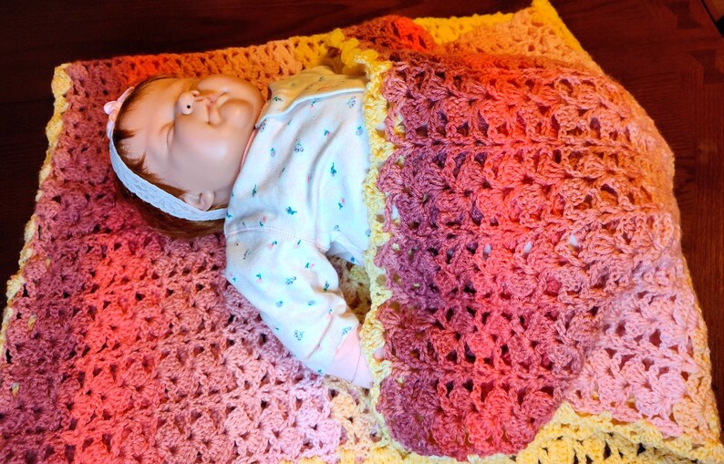 A doll is laying on it's back on a multi-colored striped blanket and is covered up to it's chest by the bottom portion of the blanket.