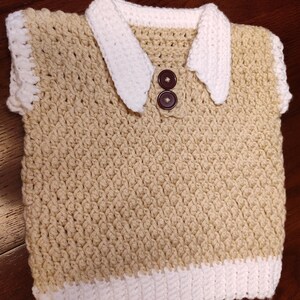 A handmade crochet vest in tan with white trim and collar. The vest features a collared neckline with two brown buttons. The bottom and edges are trimmed in white. This item is perfect for a baby or toddler.