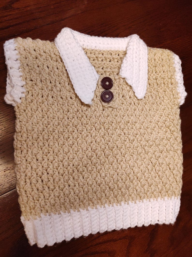 A handmade crochet vest in tan with white trim and collar. The vest features a collared neckline with two brown buttons. The bottom and edges are trimmed in white. This item is perfect for a baby or toddler.
