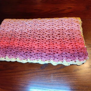 A handmade crochet blanket with a gradient of colors, transitioning from light pink to deep purple. The blanket has a scalloped yellow border, adding a decorative touch to the soft, textured surface. This item is perfect for a baby.