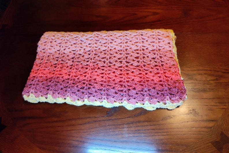 A handmade crochet blanket with a gradient of colors, transitioning from light pink to deep purple. The blanket has a scalloped yellow border, adding a decorative touch to the soft, textured surface. This item is perfect for a baby.