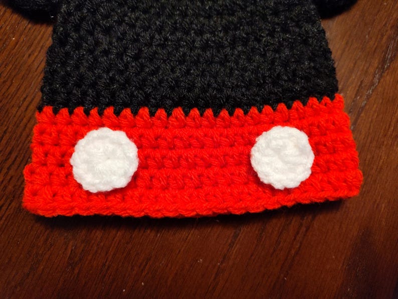 A crocheted item featuring a black upper section and a red lower section. The red portion is accented with two circular white crocheted patches positioned side by side, creating a distinct contrast against the vibrant red yarn base.
