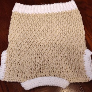 Handmade crochet sweater vest in a neutral beige color with white trim. The vest features a textured stitch pattern and a curved white waistband. The edges are finished with a white crochet border.
