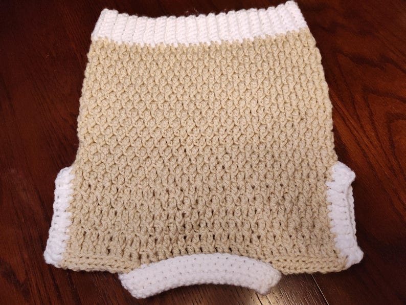Handmade crochet sweater vest in a neutral beige color with white trim. The vest features a textured stitch pattern and a curved white waistband. The edges are finished with a white crochet border.