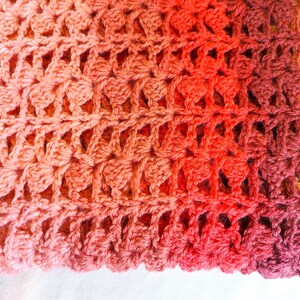 A handmade crochet blanket in shades of yellows, pinks, reds and purples. The blanket features a textured pattern and a scalloped edge.