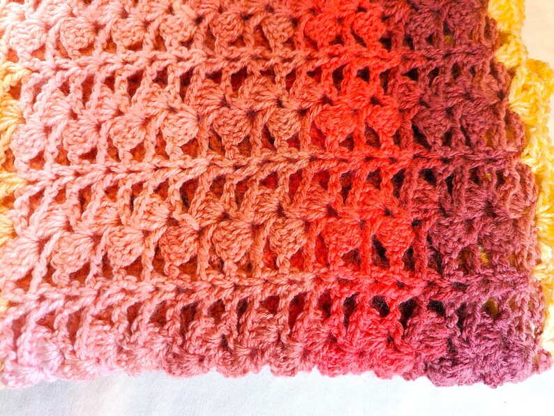 A handmade crochet blanket in shades of yellows, pinks, reds and purples. The blanket features a textured pattern and a scalloped edge.