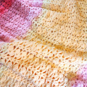 A handmade crochet blanket in shades of yellow, pink, and purple. The blanket features a textured pattern and a scalloped edge.