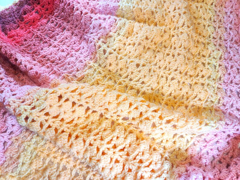 A handmade crochet blanket in shades of yellow, pink, and purple. The blanket features a textured pattern and a scalloped edge.