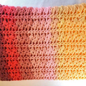 A handmade crochet blanket in shades of yellows, pinks, reds and purples. The blanket features a textured pattern and a scalloped edge.