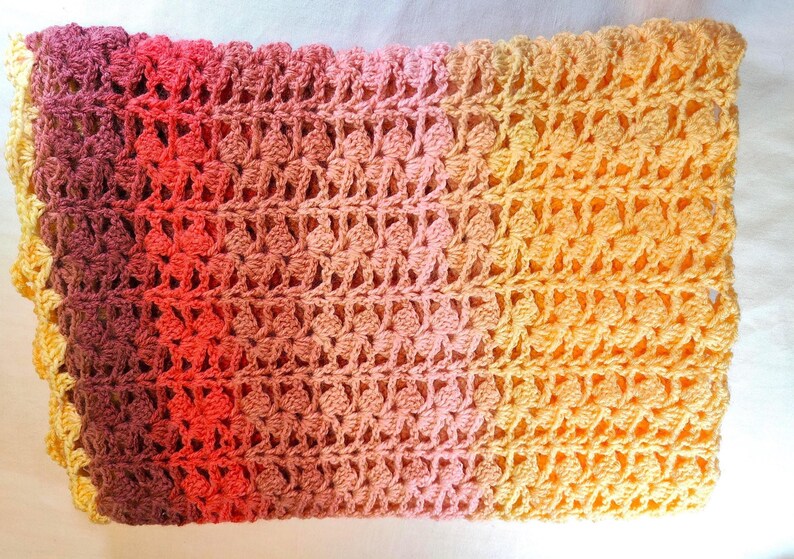 A handmade crochet blanket in shades of yellows, pinks, reds and purples. The blanket features a textured pattern and a scalloped edge.