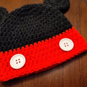 A black and red crocheted hat featuring two round black ears on top and two large white buttons attached to the front red section. The hat is constructed with a textured yarn stitch pattern and has a distinct color-blocked design.