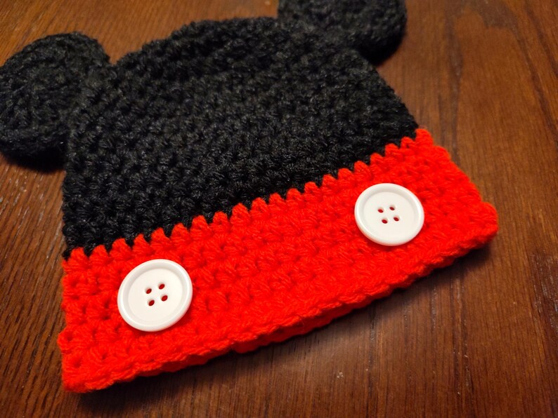 A black and red crocheted hat featuring two round black ears on top and two large white buttons attached to the front red section. The hat is constructed with a textured yarn stitch pattern and has a distinct color-blocked design.