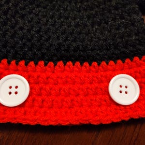 A crocheted item featuring a black upper section and a red lower section. Two large, round, white four-hole buttons are attached horizontally across the red portion of the textured yarn fabric, creating a distinct contrast.