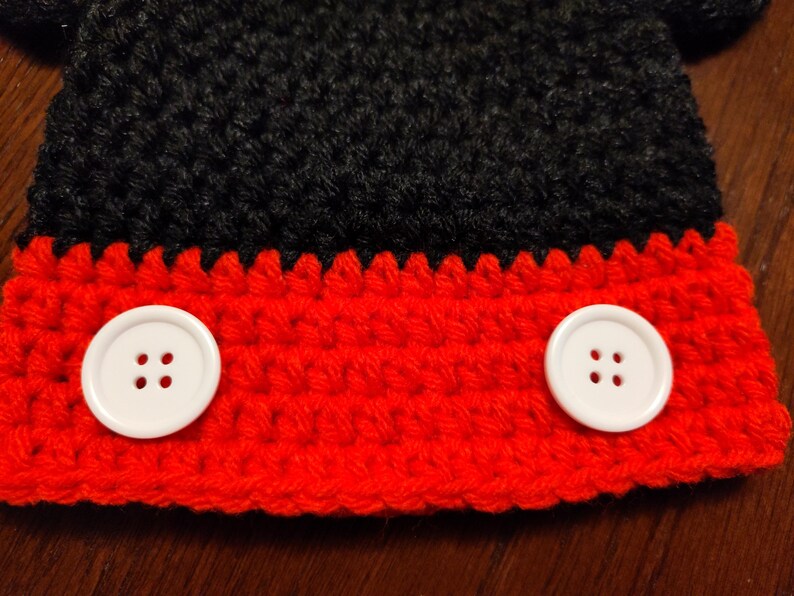 A crocheted item featuring a black upper section and a red lower section. Two large, round, white four-hole buttons are attached horizontally across the red portion of the textured yarn fabric, creating a distinct contrast.