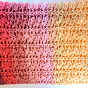 A handmade crochet blanket in shades of yellows, pinks, reds and purples. The blanket features a textured pattern and a scalloped edge.