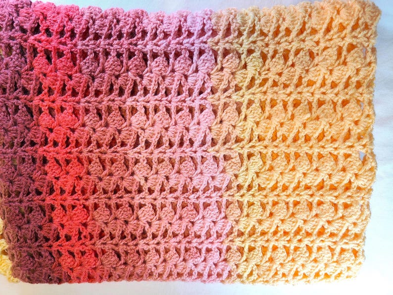 A handmade crochet blanket in shades of yellows, pinks, reds and purples. The blanket features a textured pattern and a scalloped edge.
