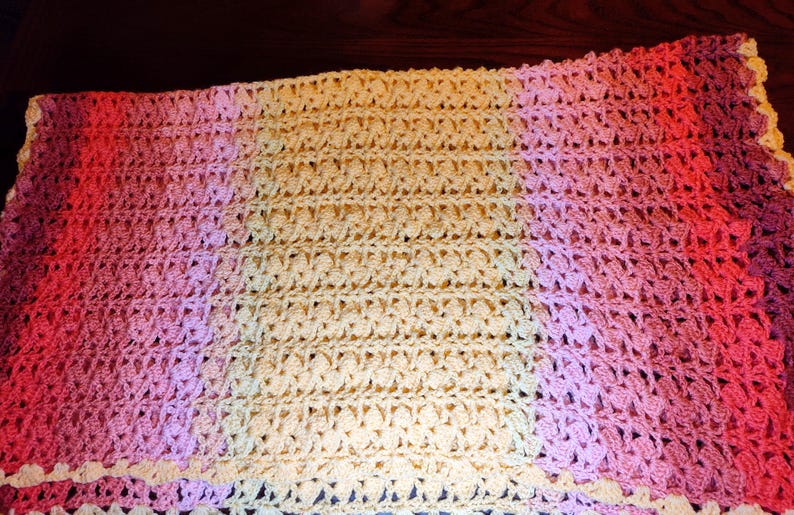 A multi-colored striped blanked is laying on a wooden table and is folded over once in half displaying a center section of yellow and end stripes of pinks, and reds.