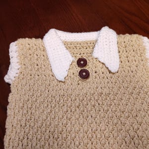 A handmade crochet top in a light tan color with a white collar and sleeve trim. Two brown buttons fasten the collar. The textured stitch creates a detailed pattern. This garment is suitable for babies or toddlers.