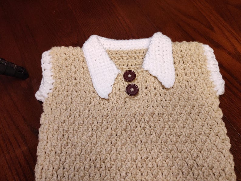 A handmade crochet top in a light tan color with a white collar and sleeve trim. Two brown buttons fasten the collar. The textured stitch creates a detailed pattern. This garment is suitable for babies or toddlers.