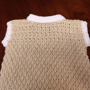 A tan crochet vest with a white collar and trim. The handmade vest features a textured stitch pattern and a classic collared neckline. The neutral color palette makes it a versatile garment for layering.