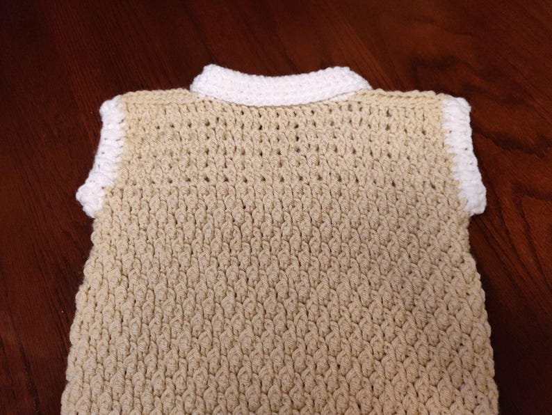 A tan crochet vest with a white collar and trim. The handmade vest features a textured stitch pattern and a classic collared neckline. The neutral color palette makes it a versatile garment for layering.