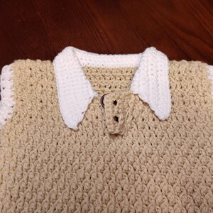 A handmade crochet top in a light tan color with a white collar and trim. The top features a textured stitch pattern and a button closure at the neckline. The collar is a contrasting white, adding a classic touch to the design.