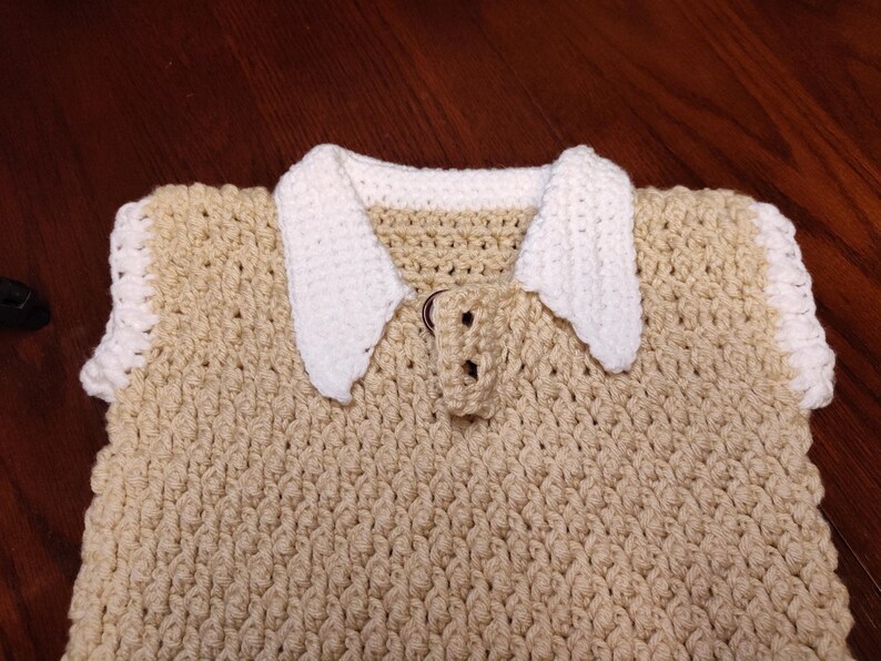 A handmade crochet top in a light tan color with a white collar and trim. The top features a textured stitch pattern and a button closure at the neckline. The collar is a contrasting white, adding a classic touch to the design.