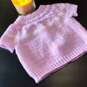A short-sleeved knit sweater in a soft lavender color. The garment features a ribbed crew neckline, a textured yoke with a subtle white horizontal stripe pattern, and a ribbed hem. The knit construction shows a consistent stitch pattern.