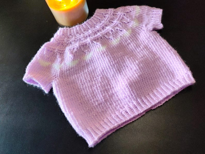 A short-sleeved knit sweater in a soft lavender color. The garment features a ribbed crew neckline, a textured yoke with a subtle white horizontal stripe pattern, and a ribbed hem. The knit construction shows a consistent stitch pattern.