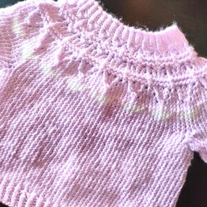 A short-sleeved, light purple knitted sweater featuring a textured yoke with a decorative pattern around the neckline. The garment is constructed with a ribbed collar, ribbed sleeve cuffs, and a ribbed hem, showcasing a consistent knit stitch throughout the body.