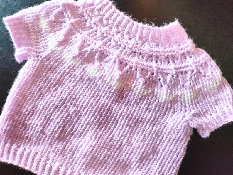 A short-sleeved, light purple knitted sweater featuring a textured yoke with a decorative pattern around the neckline. The garment is constructed with a ribbed collar, ribbed sleeve cuffs, and a ribbed hem, showcasing a consistent knit stitch throughout the body.