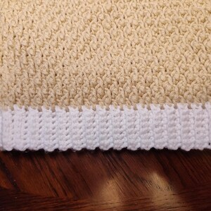 Close-up of a crocheted sweater vest with a white hem and a light beige body. The crochet stitches create a textured pattern. The vest is likely handmade and suitable for a child.