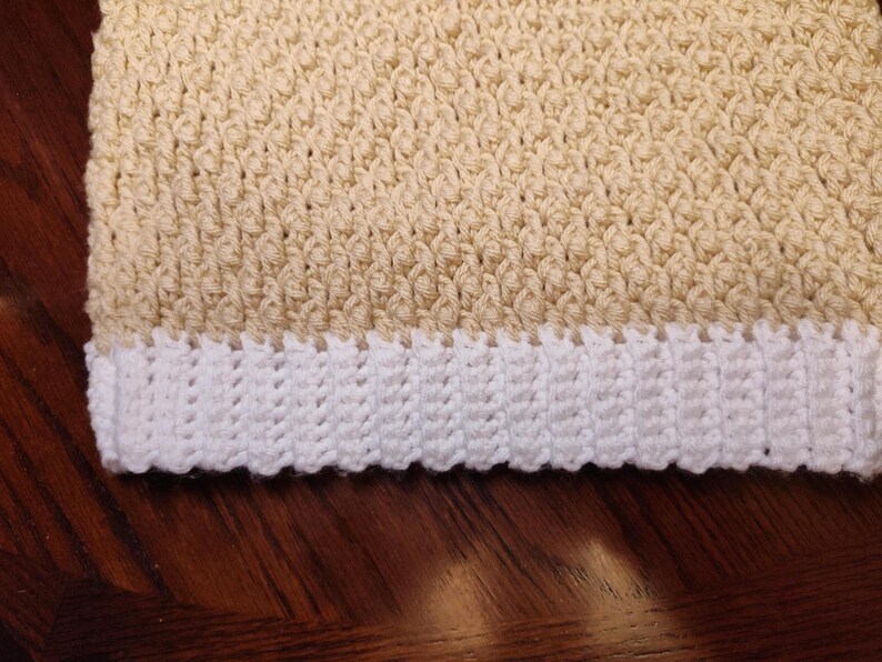 Close-up of a crocheted sweater vest with a white hem and a light beige body. The crochet stitches create a textured pattern. The vest is likely handmade and suitable for a child.