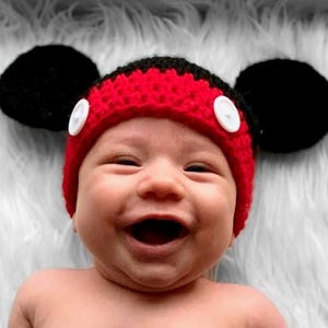 A red and black crocheted hat featuring two large round black ears on the sides and two white circular buttons attached to the front red band. The hat is constructed with a textured yarn stitch pattern and has a rounded crown shape.