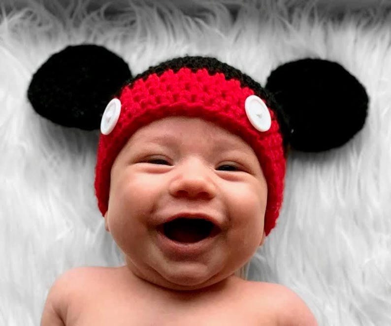 A red and black crocheted hat featuring two large round black ears on the sides and two white circular buttons attached to the front red band. The hat is constructed with a textured yarn stitch pattern and has a rounded crown shape.